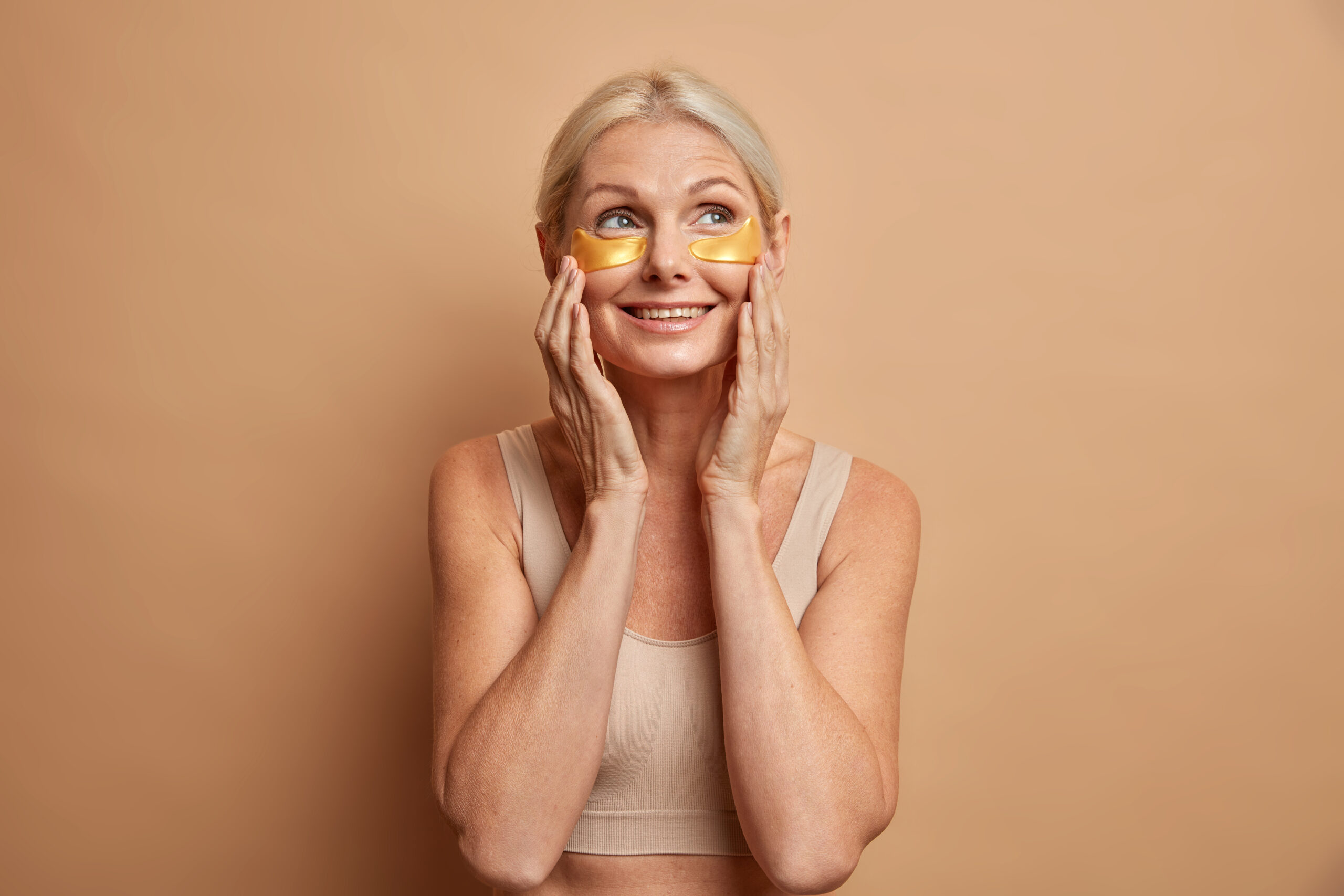 Waist up shot of glad middle aged blonde woman touches face gently applies collagen beauty patches under eyes has dreamy expression wears top poses against brown background. Facial care concept